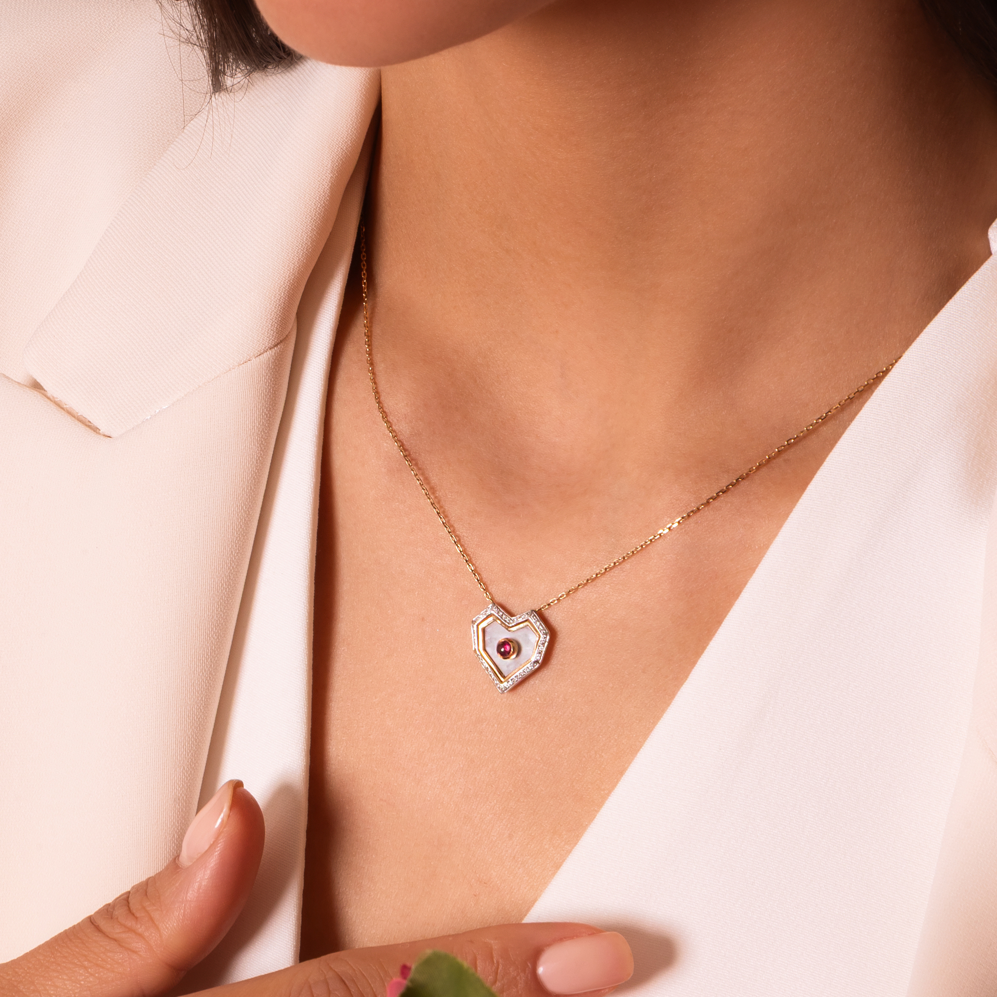 A close-up lifestyle shot of a model wearing a delicate gold heart-shaped pendant necklace with a central red gemstone, styled with a white blazer and a pink rose.