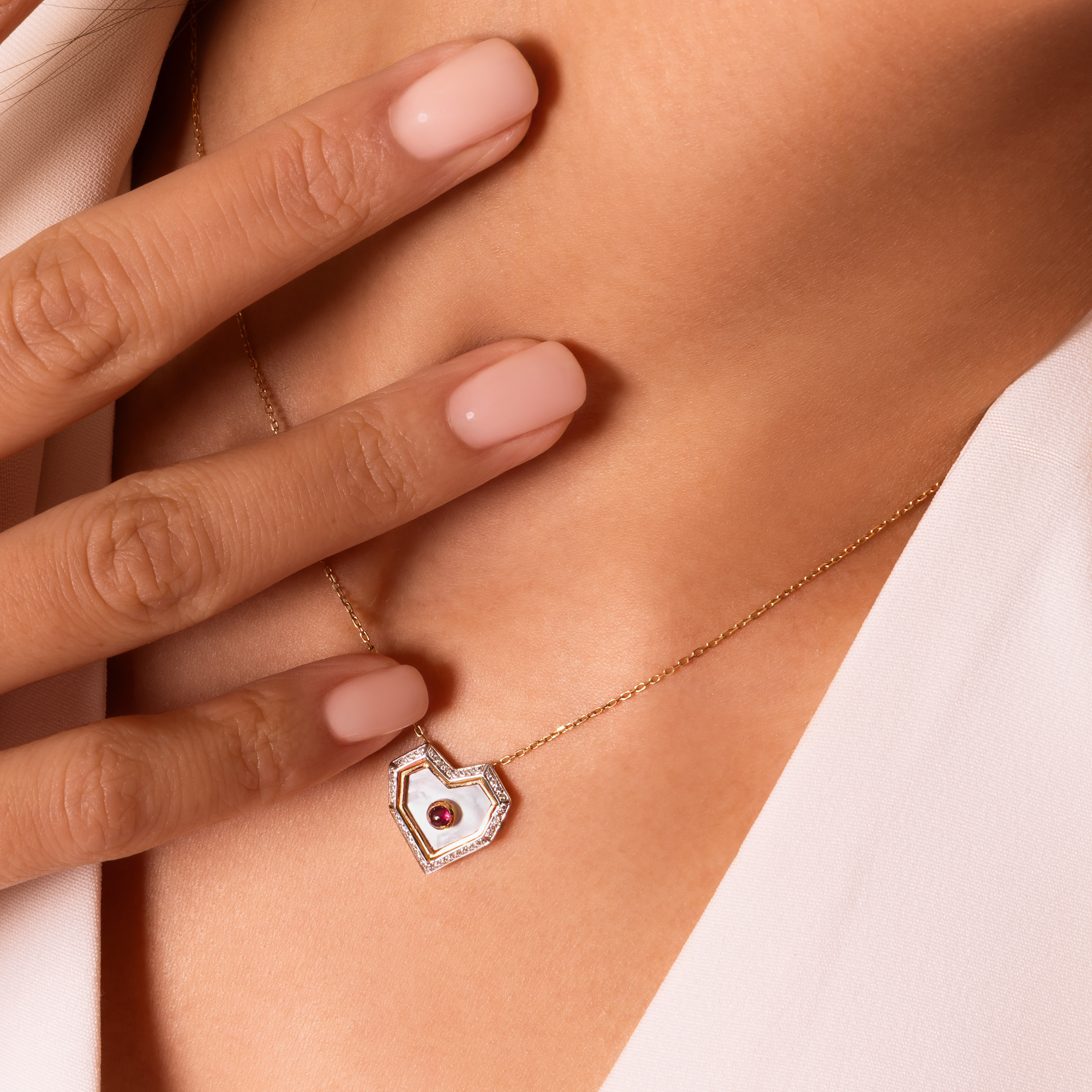 A close-up of a model's neck and hand wearing a delicate gold necklace with a shield-shaped pendant featuring a white inlay and a central red gemstone.